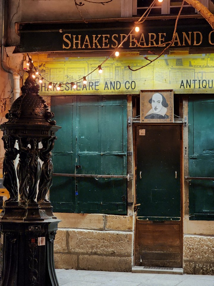 A small shop-front with green shutters closed across it, and a picture of Shakespeare over the door, with a drinking fountain in the design of four women holding up a domed roof in the left foreground.