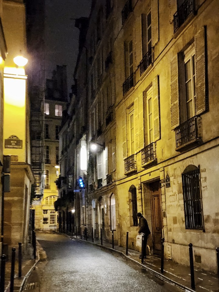 A view down a narrow street with tall townhouses leaning into it from the right, with white shutters over their windows.
