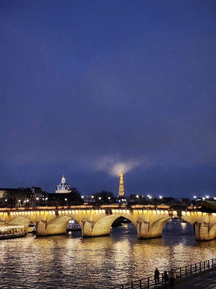 View of a bridge crossing the Seine , its street lights reflecting in the water below, with the Eiffel Tower visible in the distance, brightly lit and creating a point of light in the clouds.