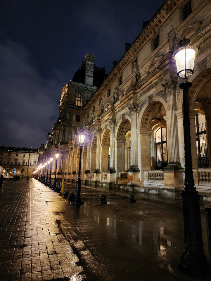 A colonnade at the Louvre, with a row of historic looking street lamps running along it, under a dark, cloudy sky.