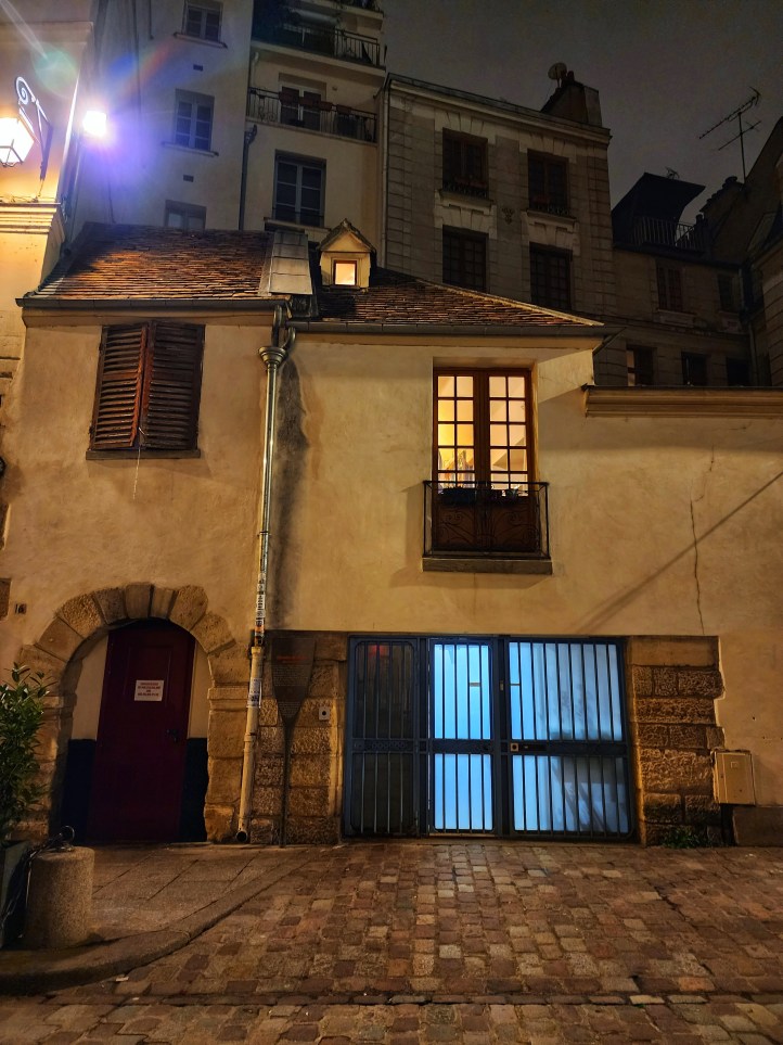 An older looking building with a rounded doorway, one small gable in the roof with orange light streaming from it, a wooden shuttered window, and a Juliet balcony with French windows. The street in front is cobbled.
