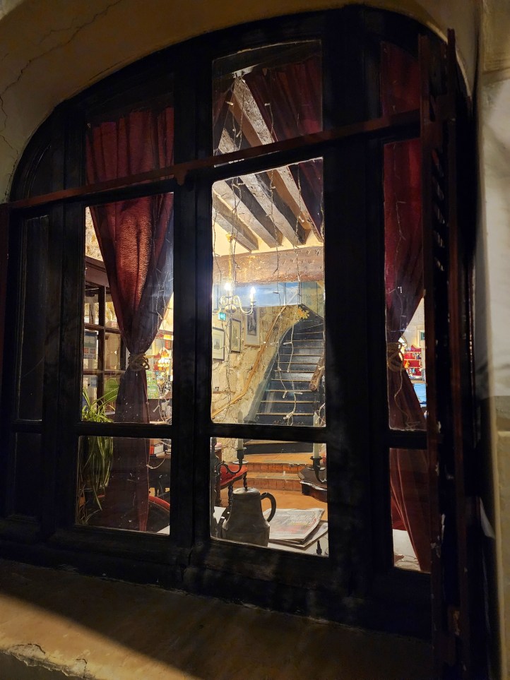 A view through a rounded, curtained window, to a wooden beamed room and a wooden, winding staircase at the back.