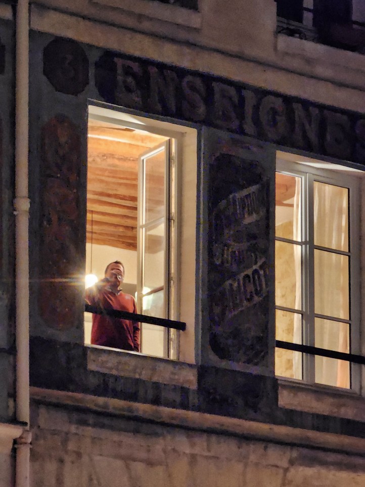 A man standing at his open window drinking a glass of wine, with ghost signs around the window.