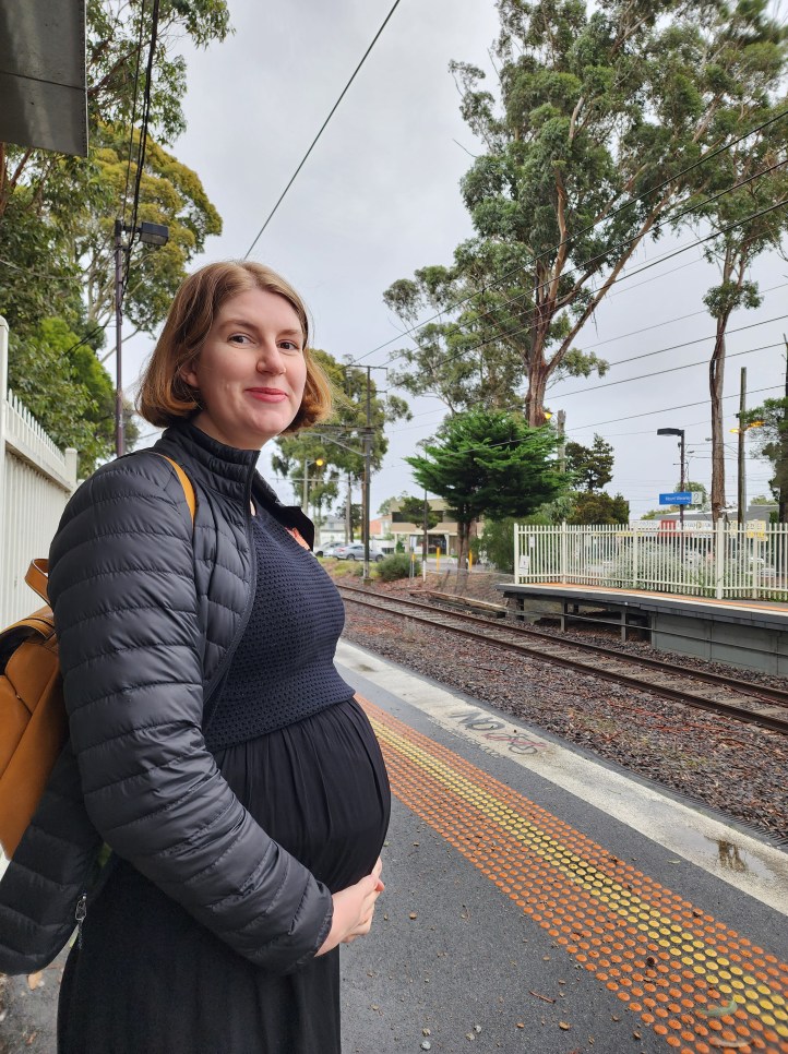 Photo of Helen in a black dress and jacket on a train station platform, with a prominent pregnancy bump.