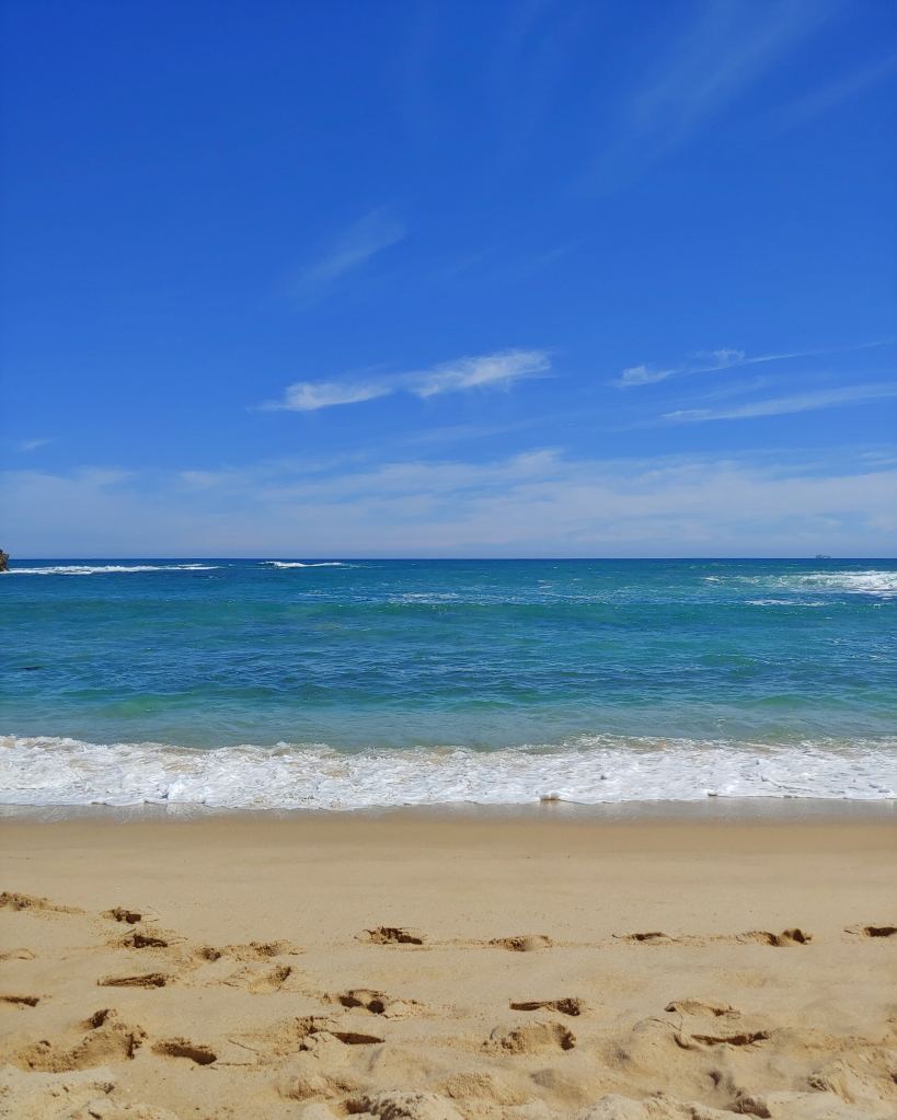 A view of a multi-coloured blue sea and gentle waves breaking on a golden sandy beach, under a bright blue sky.