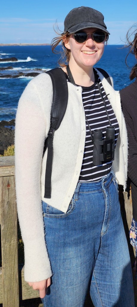 Helen wearing a cream cardigan over a black and white stripey top and blue jeans, with a pair of binoculars, round sunglasses, and a black baseball cap.