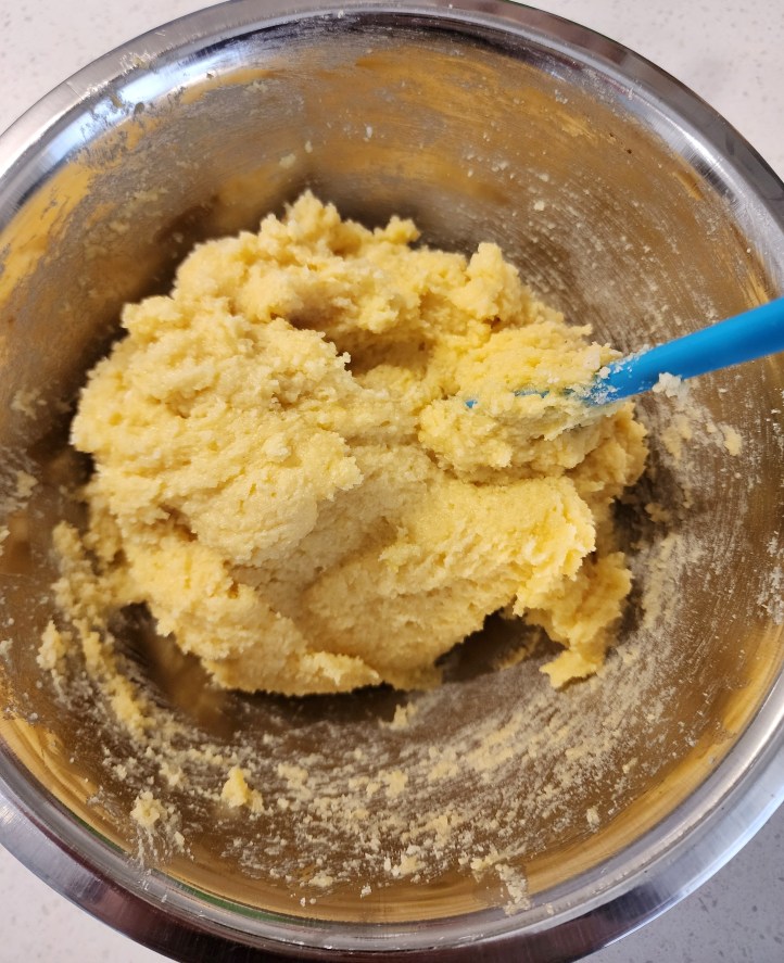 Top-down view of a batch of creamy-yellow coloured frangipane in a chrome bowl, with a small blue spatula.