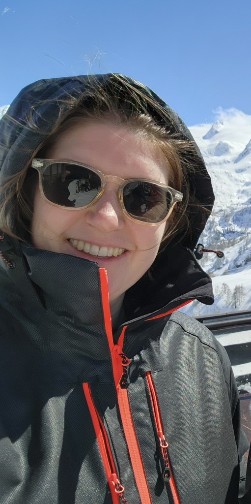 Helen wearing a black ski jacket with orange zips with the hood up, and sunglasses, against a snowy mountain backdrop.