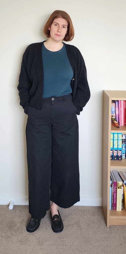 Helen wearing black wide trousers with a green/blue tight t-shirt, a black chunky cardigan open, and black loafers.