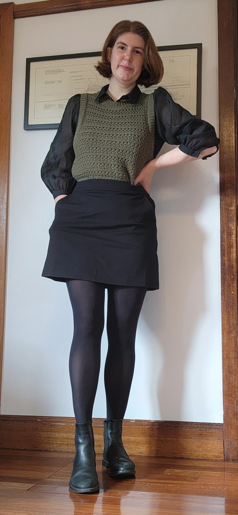 Full length photo of Helen wearing a black A-line mini skirt with a black sheer sleeved shirt, green knitted vest, and black ankle boots.