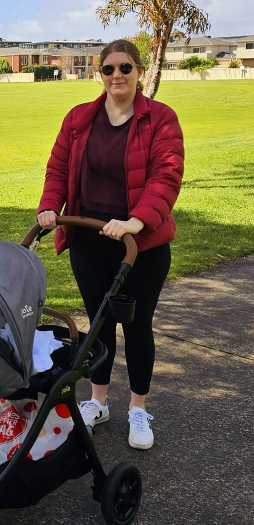 Helen wearing black maternity leggings, black and white Vejas, a burgundy sweatshirt, a red puffer jacket, and sunglasses, with her hair tied back. She is holding a pram.