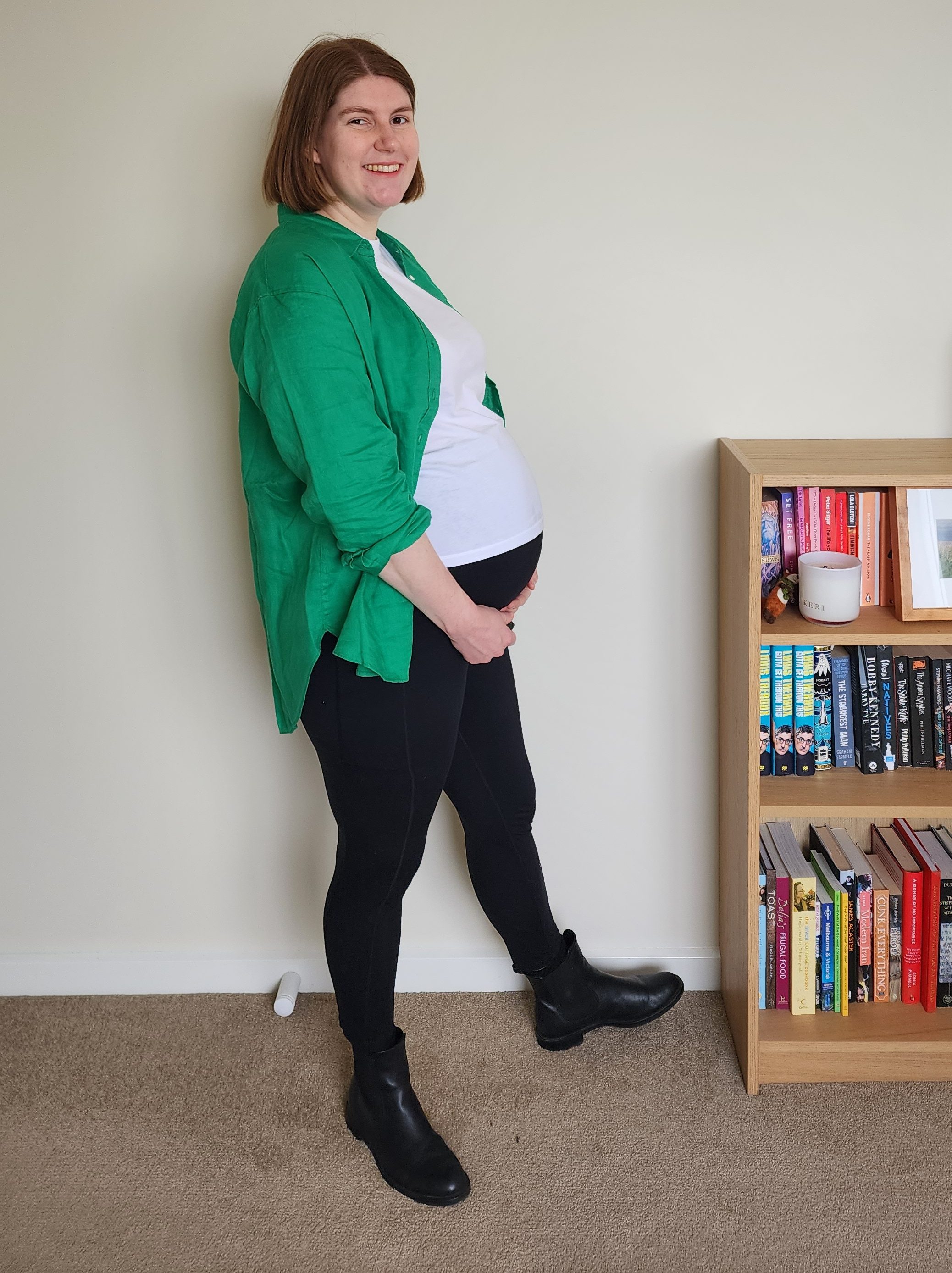 Helen wearing a white t-shirt under an open green long-sleeve linen shirt, with black maternity leggings and black chelsea boots, showing a large pregnancy bump in profile.